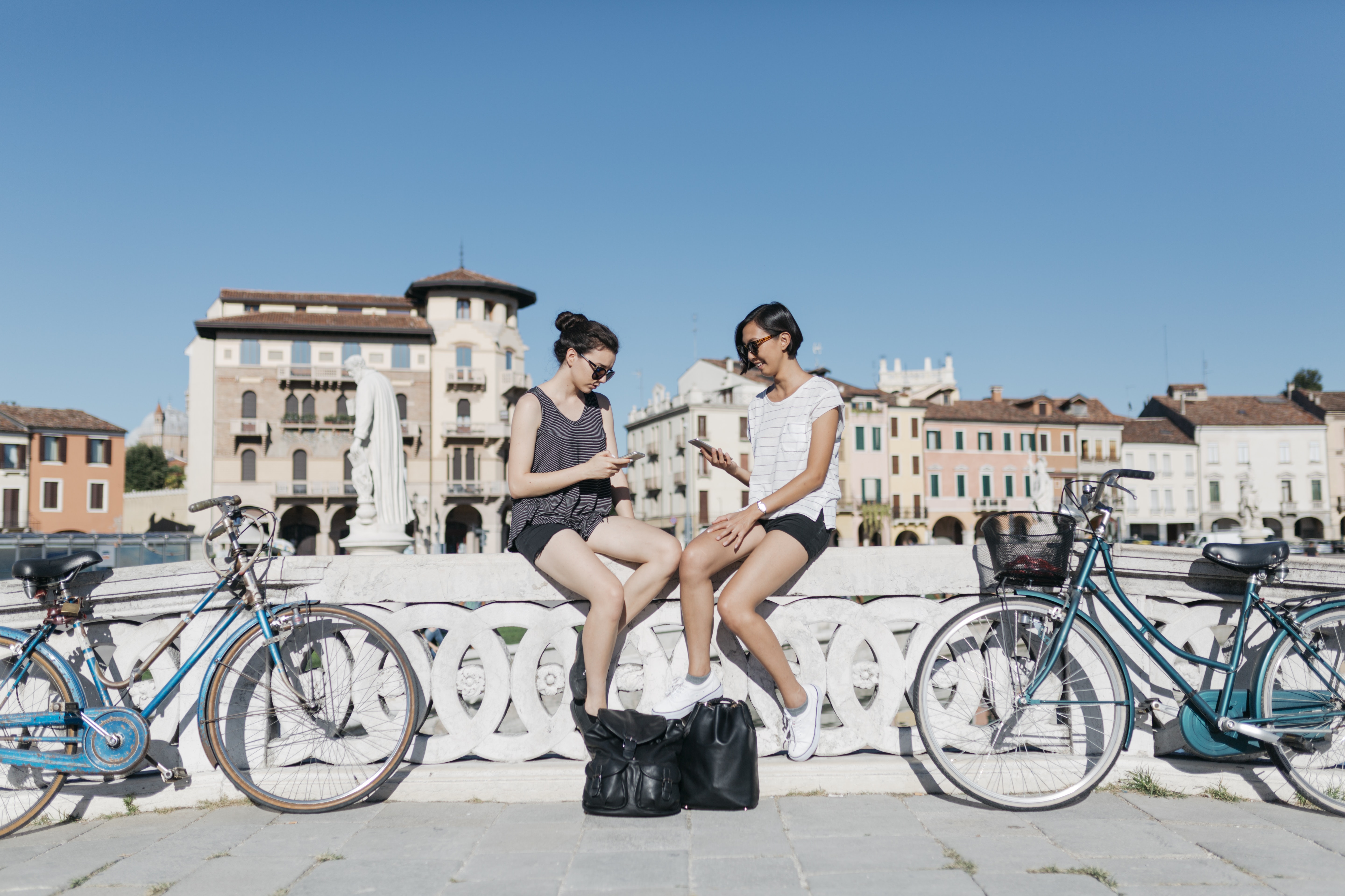 Italy, Padua, two young tourists sitting on railing looking at cell phones